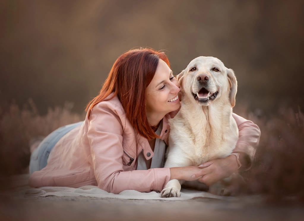 How to preserve your pet's memory through photography in san diego 3 Red-haired woman lying in a soft, neutral-toned outdoor setting while hugging her Labrador Retriever, both radiating warmth and love. Emotional pet photography by Kat De Laet, San Diego.