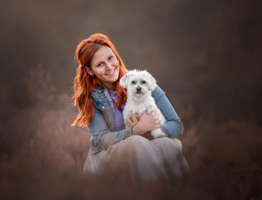 Woman in a flowing blue dress holding a small white dog, both posing elegantly against a softly blurred background. High-end pet and owner photography by Kat De Laet, San Diego.