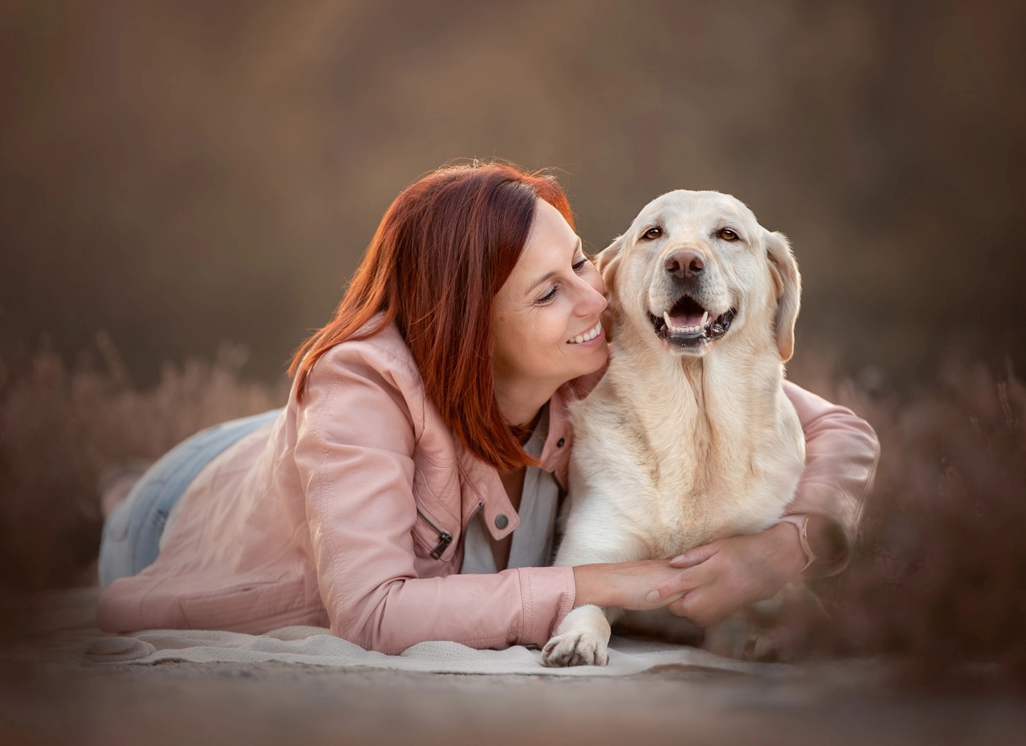 10 tips to help your dog stay calm during a photo session in san diego 2 Red-haired woman lying in a soft, neutral-toned outdoor setting while hugging her Labrador Retriever, both radiating warmth and love. Emotional pet photography by Kat De Laet, San Diego.