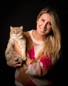 Woman-holding-british-shorthair-cat Blonde woman in a pink and white sweater holding a golden British Shorthair cat, both smiling toward the camera. Elegant pet portrait photography by Kat De Laet, San Diego.