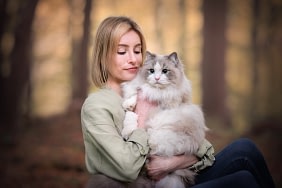 Woman-holding-fluffy-cat-in-forest Young woman in a green blouse gently cradling a Siberian cat with soft golden light filtering through the trees. Fine art pet portrait by Kat De Laet, San Diego.