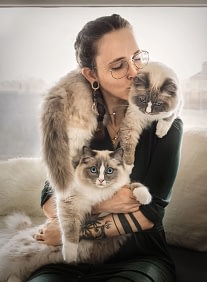 Woman-holding-two-fluffy-ragdoll-cats Tattooed woman holding two Ragdoll cats, both looking toward the camera with a cozy, intimate feel. Artistic pet photography by Kat De Laet, San Diego.