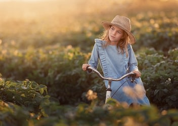 Girl-in-hat-on-bicycle-field Young girl in a straw hat and denim dress riding a vintage bicycle through a lush green field at sunset. Dreamy fine art children's photography by Kat De Laet, San Diego.