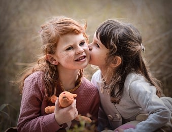 Kids-sharing-wholesome-moment-outdoors family photo session Two young girls sharing a sweet moment, one whispering into the other's ear. Heartfelt childhood portrait photography by Kat De Laet, San Diego.