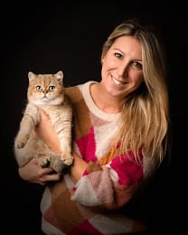 Woman-holding-british-shorthair-cat Blonde woman in a pink and white sweater holding a golden British Shorthair cat, both smiling toward the camera. Elegant pet portrait photography by Kat De Laet, San Diego.