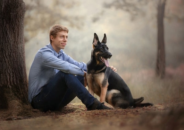 Man-german-shepherd-tree family photo session Man sitting with his German Shepherd in a peaceful autumn park, both looking contemplative. Fine art dog photography by Kat De Laet, San Diego.