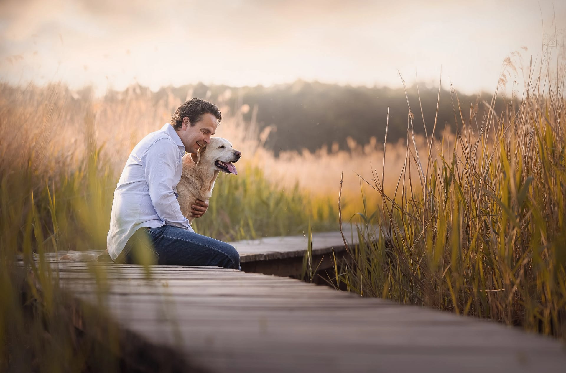 Heartfelt moment between a man and his Labrador Retriever, embracing on a wooden path in a golden field at sunset. Emotional pet portrait by Kat De Laet in San Diego.