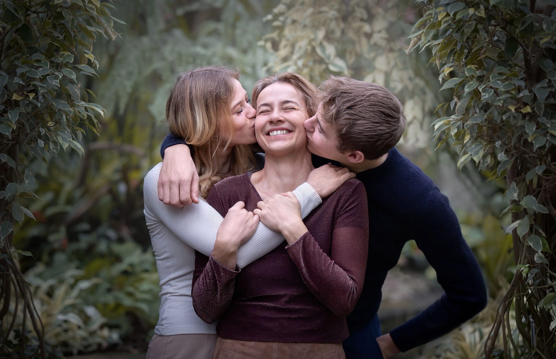 Mother laughing as her two teenage children kiss her cheeks, surrounded by lush greenery. Heartwarming family photography by Kat De Laet, San Diego.