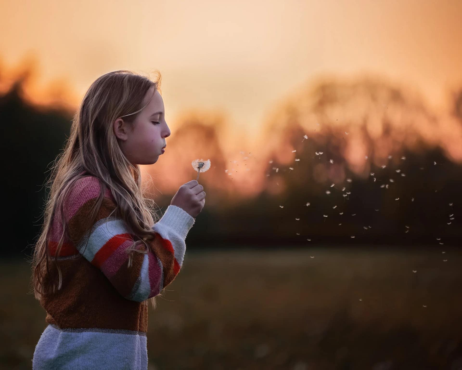Young girl making a wish while blowing on a dandelion at sunset, with warm golden light in the background. Fine art children's photography by Kat De Laet, San Diego.
