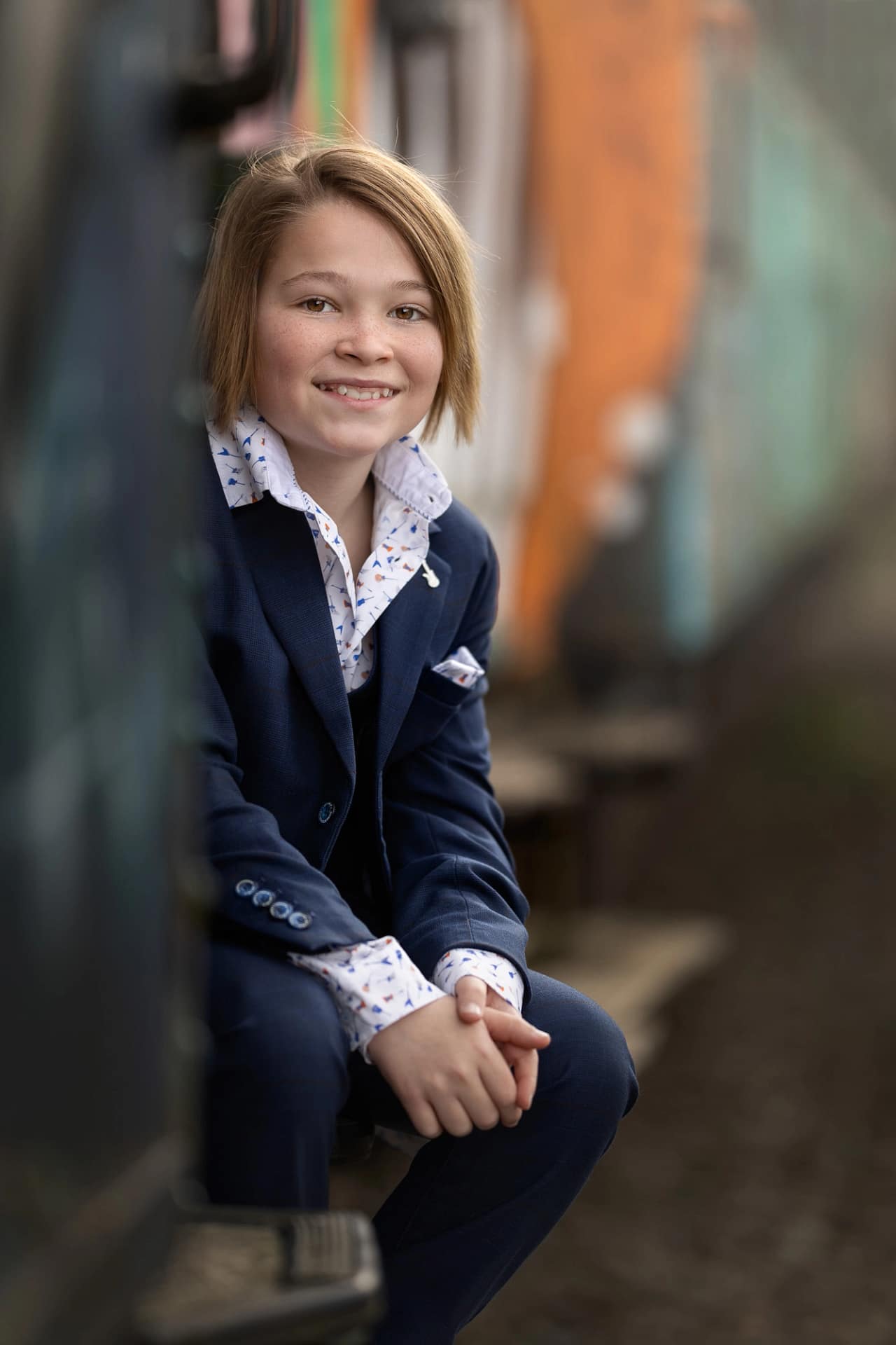 Smiling boy in a tailored suit sitting outdoors in a rustic alleyway. Stylish children’s portrait by Kat De Laet, San Diego.