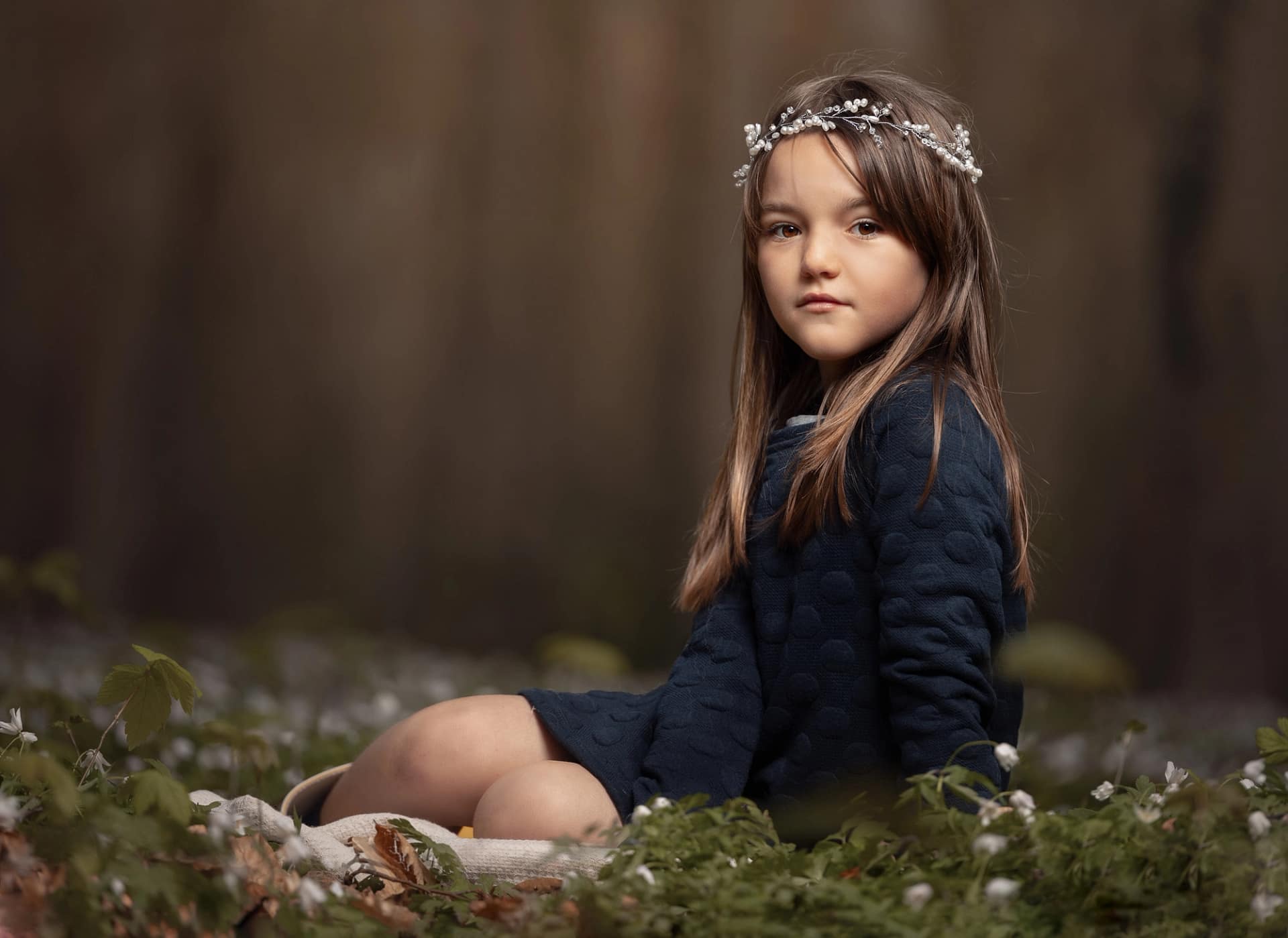 Young girl in a dark dress sitting on a mossy log with a floral headband, gazing softly. Fine art children's photography by Kat De Laet, San Diego.