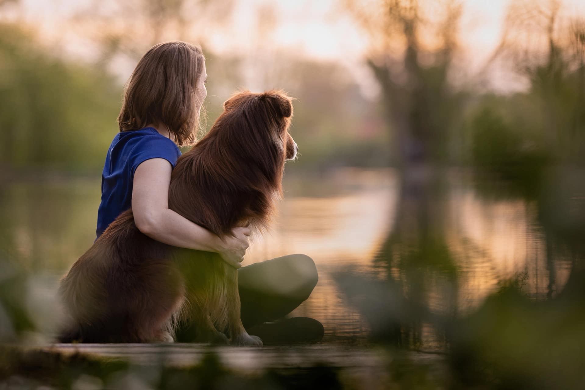 Woman sitting by a reflective lake with her long-haired dog, both gazing at the serene water during golden hour. Dreamy pet photography by Kat De Laet, San Diego.