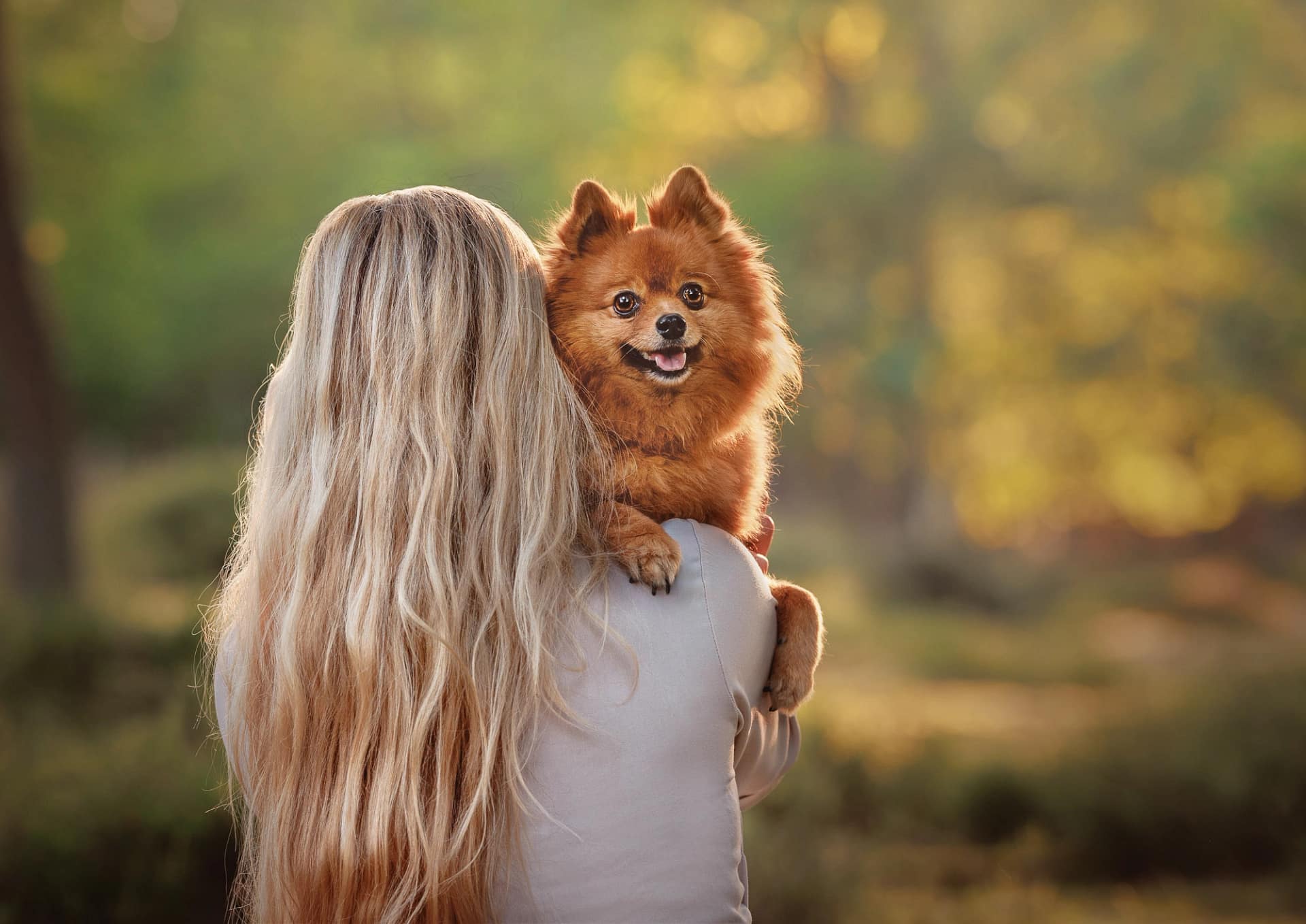 Blonde woman holding a fluffy Pomeranian over her shoulder, both bathed in warm sunset light. Luxury pet and owner photography by Kat De Laet, San Diego.
