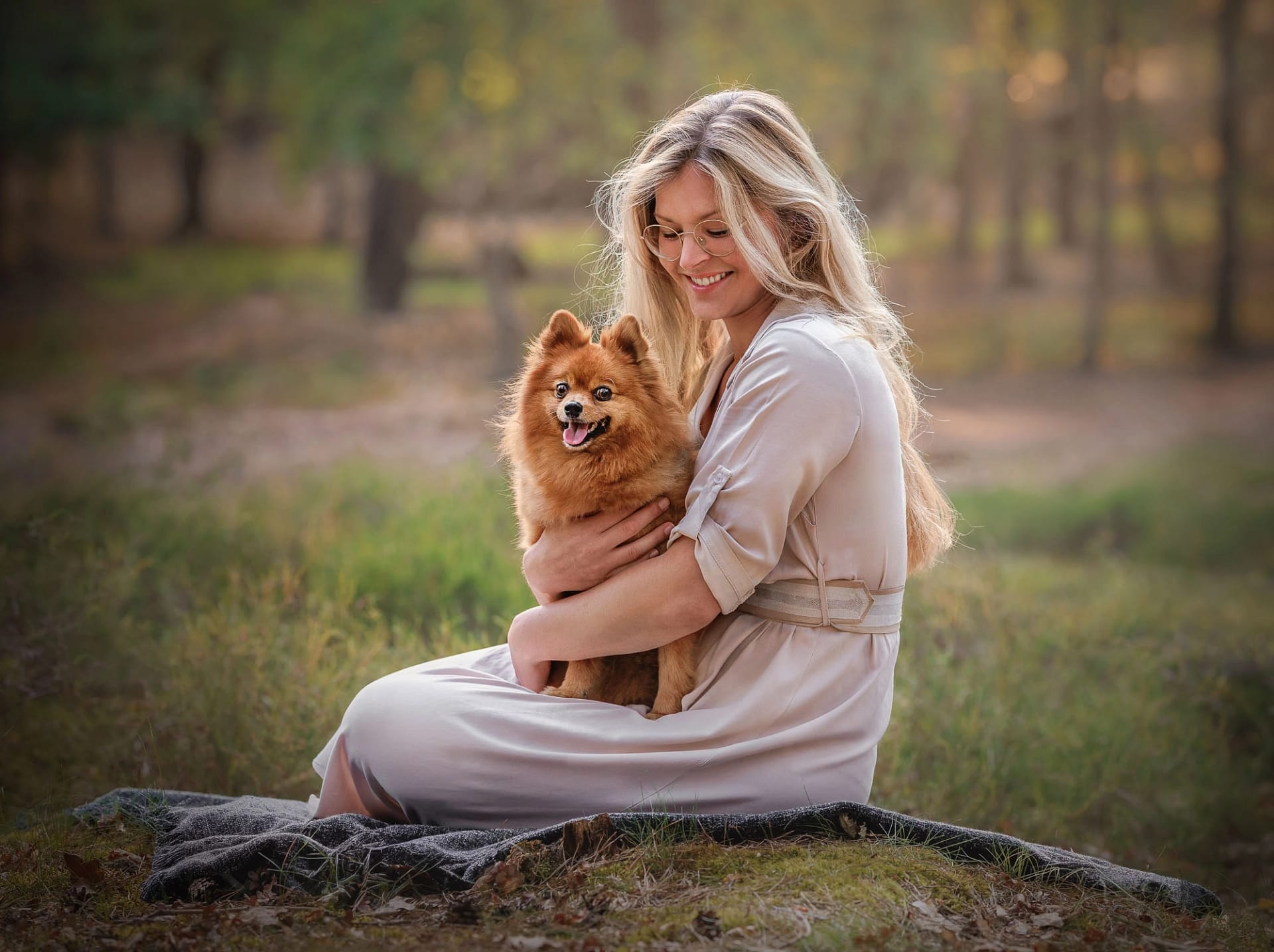 Woman in a beige dress embracing a fluffy Pomeranian dog in a forest setting.